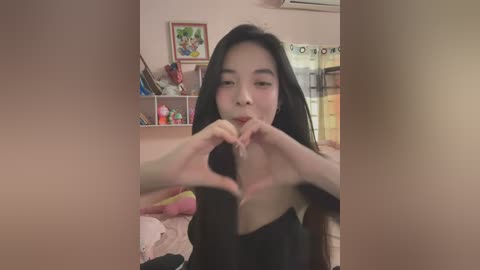 Media: Video of an Asian woman with long black hair, fair skin, and small breasts, making heart shapes with her hands in a bedroom with pink walls, stuffed toys, and shelves.