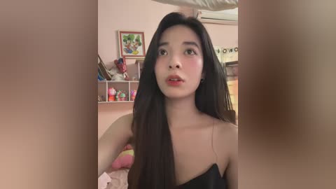 Media: Video of an Asian woman with long black hair, wearing a black strapless top, standing in a bedroom with pink walls, shelves with toys, and framed pictures.