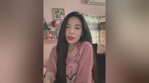 Media: Video of an East Asian woman with long black hair, wearing a pink shirt, sitting in a cozy, cluttered room with pastel walls, a window, and framed art.