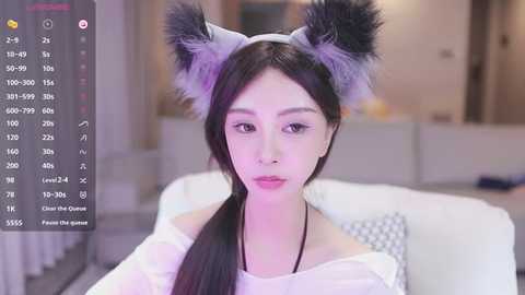 Media: A video of an East Asian woman with light skin, wearing a grey off-shoulder top and grey wolf ears, seated on a white couch in a modern, dimly lit room.