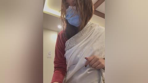 Media: Video of a person with wet hair, wearing a blue surgical mask and a red long-sleeve shirt, holding a white towel. Background features beige walls and a ceiling light.