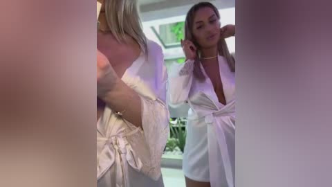 Media: Video of two women in white robes, one blonde, one brunette, talking in a modern, minimalist room with greenery outside the window.