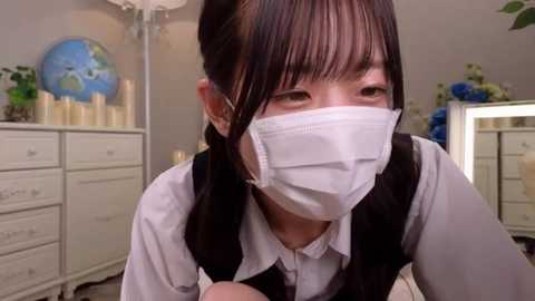 Media: Video of an Asian woman in a white mask, black vest over a white shirt, in a brightly lit room with white furniture and candles.