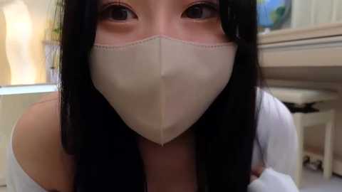 Media: A video of a young woman with long black hair, wearing a white surgical mask and a white off-shoulder top, standing indoors in a blurred kitchen with cabinets and a countertop.