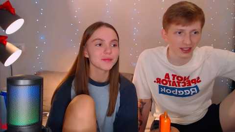 Media: Video of a young white couple with long hair, sitting on a couch in a cozy living room. The boy wears a \"Retro Pop\" shirt, and the girl a blue jacket.