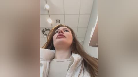 Media: Video of a young woman with long, straight auburn hair, wearing a white shirt, looking upwards. She has a serene expression. Background includes white ceiling tiles and a ceiling fan.