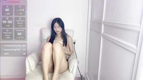 Media: Video of a slim, nude Asian woman with long black hair, sitting in a white armchair, legs crossed, in a minimalist room with a digital thermometer display.