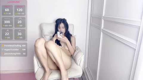 Media: Video of a naked Asian woman with black hair, sitting in a white leather chair, holding a phone, against a white wall and closet.