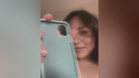 Media: Video of a woman with shoulder-length dark hair, smiling, taking a mirror selfie with a teal phone, blurry background.