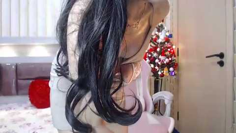 Media: Video of a woman with long black hair, wearing a white dress, bending forward, reflecting in a mirror, surrounded by a Christmas tree with ornaments, and a red chair in a cozy, festive bedroom.