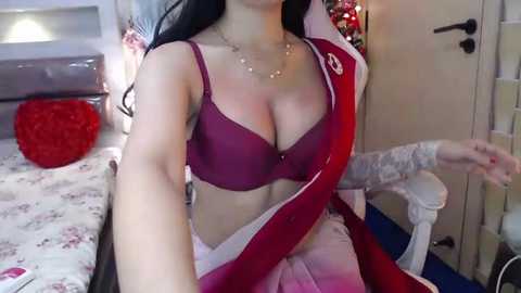 Media: Video of a woman with long black hair, wearing a maroon bra and white saree, sitting on a bed in a cozy bedroom with floral bedspread, red pillow, and Christmas decorations.