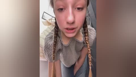 Media: A video of a young girl with fair skin, brown hair braided into pigtails, wearing a grey sweater with puffed sleeves, and a low-cut top revealing her midriff. She has a neutral expression, and the background shows a blurred indoor setting.