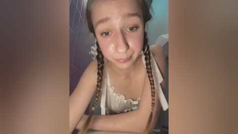 Media: A video of a young girl with light skin, brown braided pigtails, and a white tank top, sitting indoors with a slightly mischievous expression.