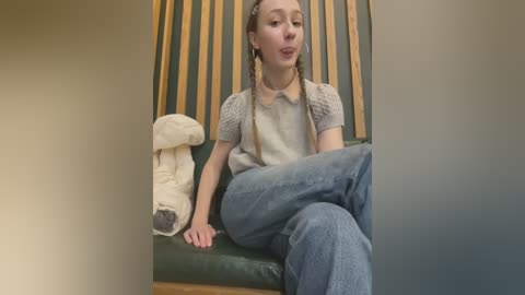 Media: Video of a young Caucasian girl with long braids, wearing a gray sweater and blue jeans, sitting on a green cushioned bench. A stuffed mushroom and striped wall backdrop are visible.