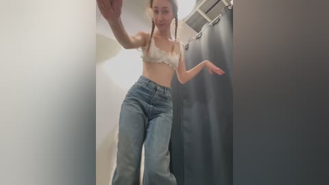 Media: Video of a young woman in a fitting room, wearing a white lace bra and high-waisted blue jeans. She stands with one arm raised, smiling, against a gray curtain.