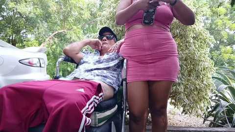 Media: Video of an older man in a wheelchair, wearing a plaid shirt and red shorts, with a young woman in a pink dress standing beside him in a lush garden.