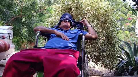 Media: Video of an elderly person with dark skin, wearing a blue shirt, red pants, and a dark hat, relaxing in a wheelchair amidst lush greenery, surrounded by trees and plants.