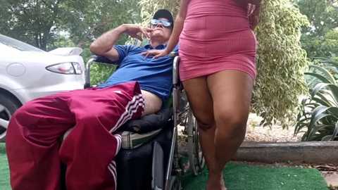 Media: A video of an older man in a wheelchair, wearing a blue shirt and red tracksuit, with a woman in a pink mini dress standing beside him.