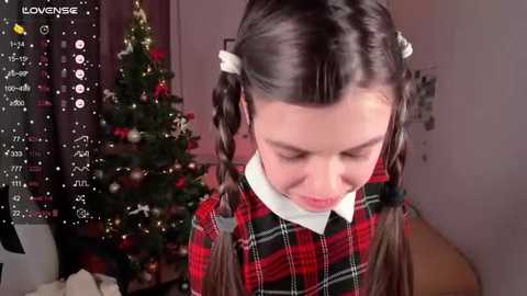 Media: A young girl with pigtails and a red plaid dress stands in a cozy, Christmas-decorated room. The background features a lit Christmas tree and warm lighting.