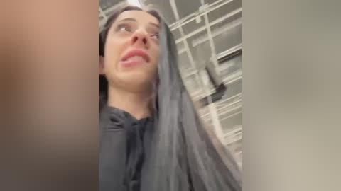 Media: Video of a young woman with long, straight, gray hair, wearing a black shirt, looking up with a surprised expression, in a bathroom with a white tiled ceiling and shower.