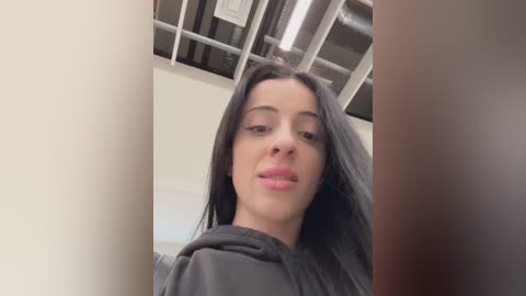 Video of a young woman with long black hair, light skin, and full lips, wearing a gray hoodie. She's indoors with a ceiling full of exposed pipes. The background is blurred.