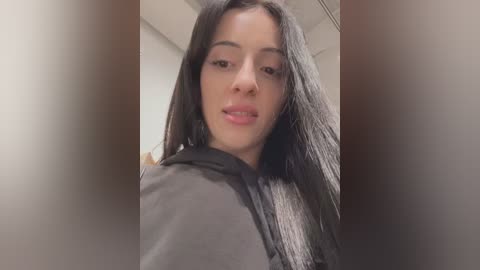 A video of a young woman with long, straight black hair, wearing a grey hoodie, captured from a low angle, emphasizing her face. The background is blurry, showing a white wall and ceiling.