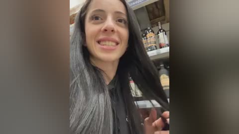 Video of a smiling woman with long, straight, dark hair in a salon, holding a bottle of hair dye, with shelves of hair products in the background.