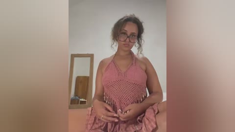 Media: A video of a curly-haired woman with glasses wearing a pink crochet top, standing in a beige room with a wooden mirror and a round brown object.