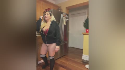 Media: Video of a curvy, blonde woman in a black jacket, red lingerie, and thigh-high black socks, posing in a cozy kitchen with wooden floors and a stainless steel fridge.