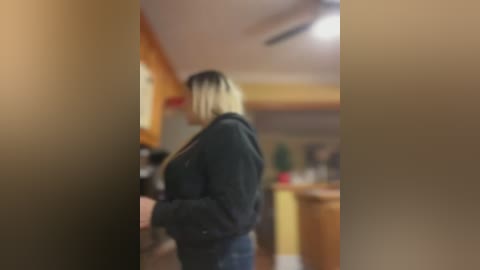 Media: A blurred video of a blonde woman in a black jacket standing in a kitchen with wooden cabinets and a stove.