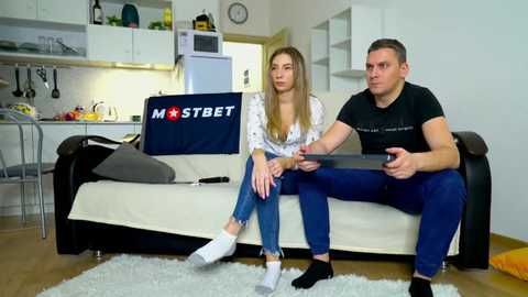 Media: Video of a young white couple sitting on a beige sofa in a modern, minimalist living room. The man, wearing a black t-shirt and blue jeans, plays a video game. The woman, in a white blouse and blue jeans, looks thoughtful.