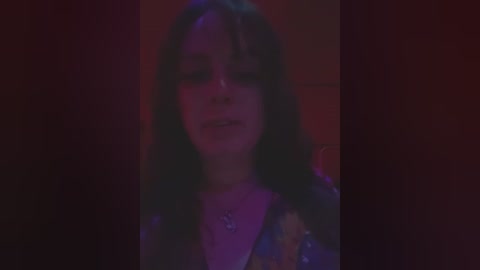 Media: A dimly-lit video of a young woman with long, dark hair and pale skin, wearing a colorful, sequined top, standing against a dark, blurred background.