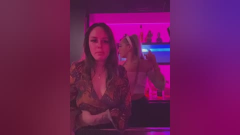 Media: Video of a woman with long brown hair, wearing a floral blouse, looking serious in a dimly lit bar with neon lights and a blurred bartender in the background.