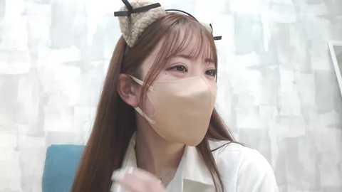 Media: Video of a young woman with long brown hair, wearing a white mask, a white lab coat, and a cat ear headband. She's in a sterile, white-walled room with a blue chair in the background.