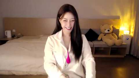 Media: Video of a smiling East Asian woman with long black hair in a white coat, sitting on a bed with a teddy bear, in a warmly lit, modern bedroom.