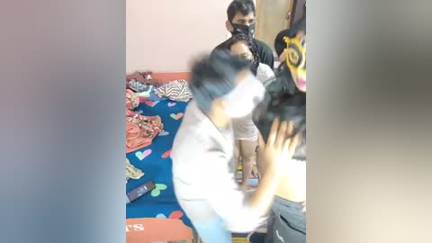 Media: A video shows a young girl in a white shirt and black pants being held by an adult man with a black mask in a cluttered room with a blue and orange bedspread.