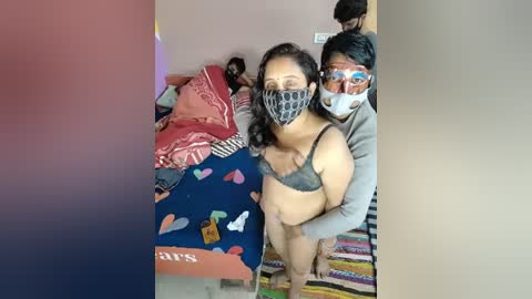 Media: Video of a woman wearing a mask and gray bra, standing in a messy room with colorful bedding, a man behind her.