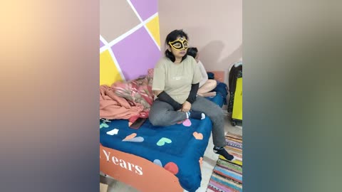 Media: A video shows a young woman with medium skin tone, wearing a yellow mask and gray sweatshirt, sitting on a blue bed adorned with \"Years\" and colorful heart-shaped pillows in a colorful, patterned room.