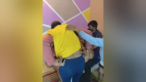 Media: Video of two men restraining a large, muscular man in a yellow vest, blue pants, and pink shirt in a room with colorful, geometric-patterned walls.