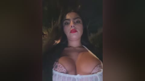 Media: A video of a voluptuous woman with long dark hair, large breasts, and fair skin, wearing a white lace bra. The background is dark and blurred, emphasizing her.