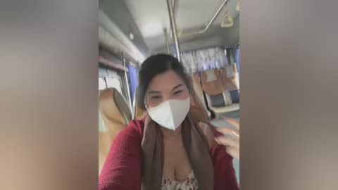 Media: Video of a woman with long black hair, wearing a red cardigan and white face mask, seated on a public bus with rows of seats and windows in the background.