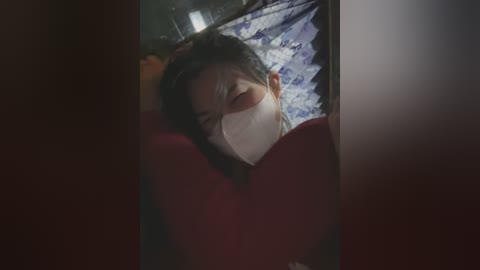 Media: A dimly lit video shows a person with light skin, dark hair, and a white face mask, sleeping in a dimly lit room with a patterned blue and white quilt.