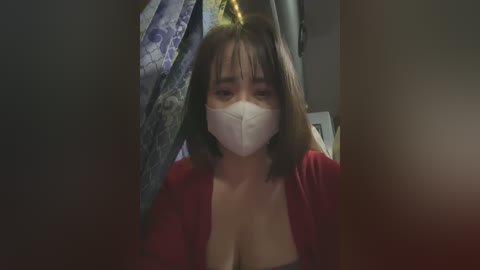 Media: Video of a young woman with shoulder-length dark hair, wearing a red cardigan, white face mask, and a low-cut top, partially obscured by a curtain, creating a mysterious and intimate atmosphere.