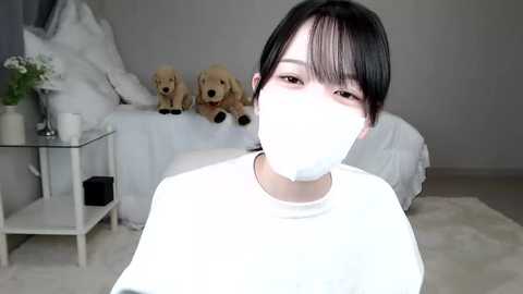 Media: Video of an East Asian woman with short black hair, wearing a white mask, white t-shirt, and a beige blanket, in a minimalist bedroom with a white bed, stuffed animals, and a small table.