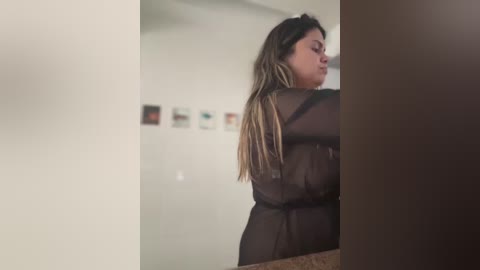 Media: Video of a woman with long, dark hair, wearing a sheer black blouse, seen from the side in a dimly lit room with blurred, framed artwork on a white wall.