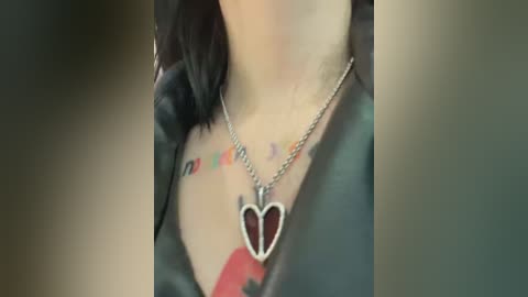 Media: A close-up video of a woman's neck and chest, adorned with a black leather jacket and a silver heart pendant necklace. Visible tattoos include a small \"love\" design and a red heart.