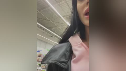 Media: A video showing a close-up of a woman with long black hair, wearing a pink top and black leather jacket, standing in a grocery store with fluorescent lighting and visible shelves.