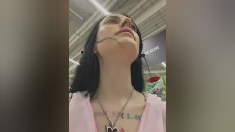 Media: A video of a young woman with fair skin and long black hair, wearing a pink cardigan, standing indoors with a tattoo on her chest, smiling and looking up.