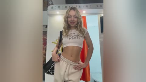 Media: Video of a blonde woman in a cropped white tank top and loose beige pants, standing confidently in a modern store with white walls, orange curtains, and \"Women's Shoes\" signage.