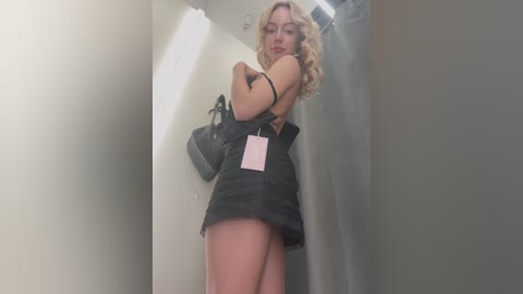 Media: Video of a blonde woman with curly hair, wearing a black mini dress, lifting it to reveal her buttocks, standing in a narrow, dimly-lit hallway.